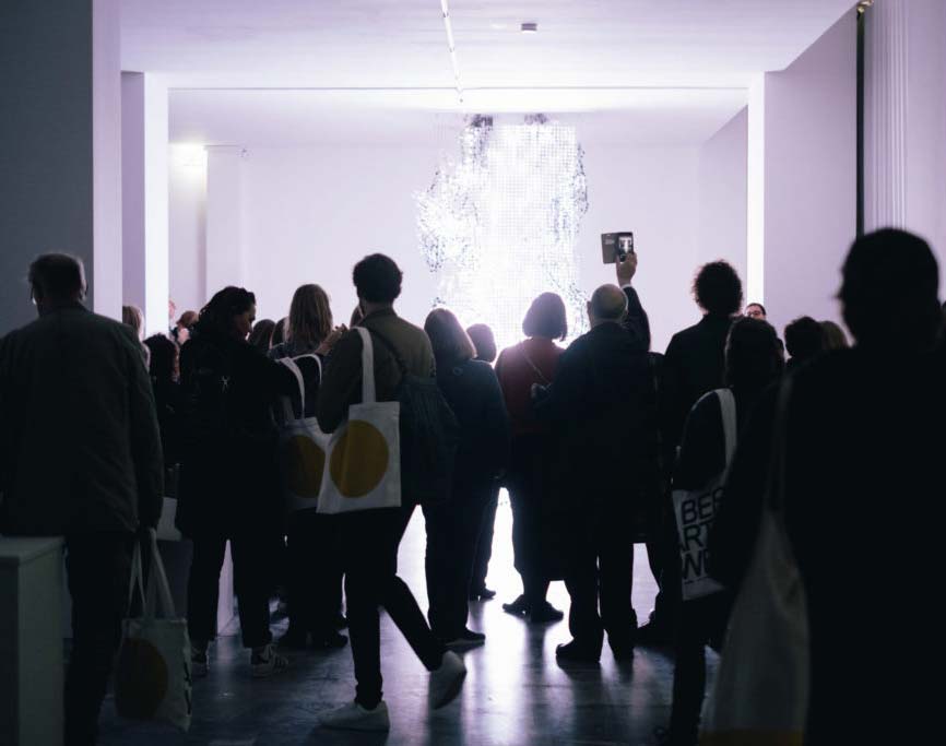 Besucher stehen in einem hellen Raum und bewundern eine Lichtinstallation.