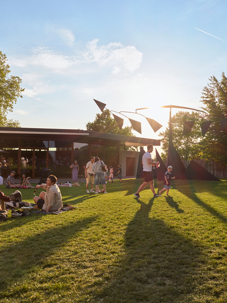 Menschen verbringen die Zeit in einer sonnigen Parklandschaft mit modernen Skulpturen und einem Caféhäuschen im Hintergrund.